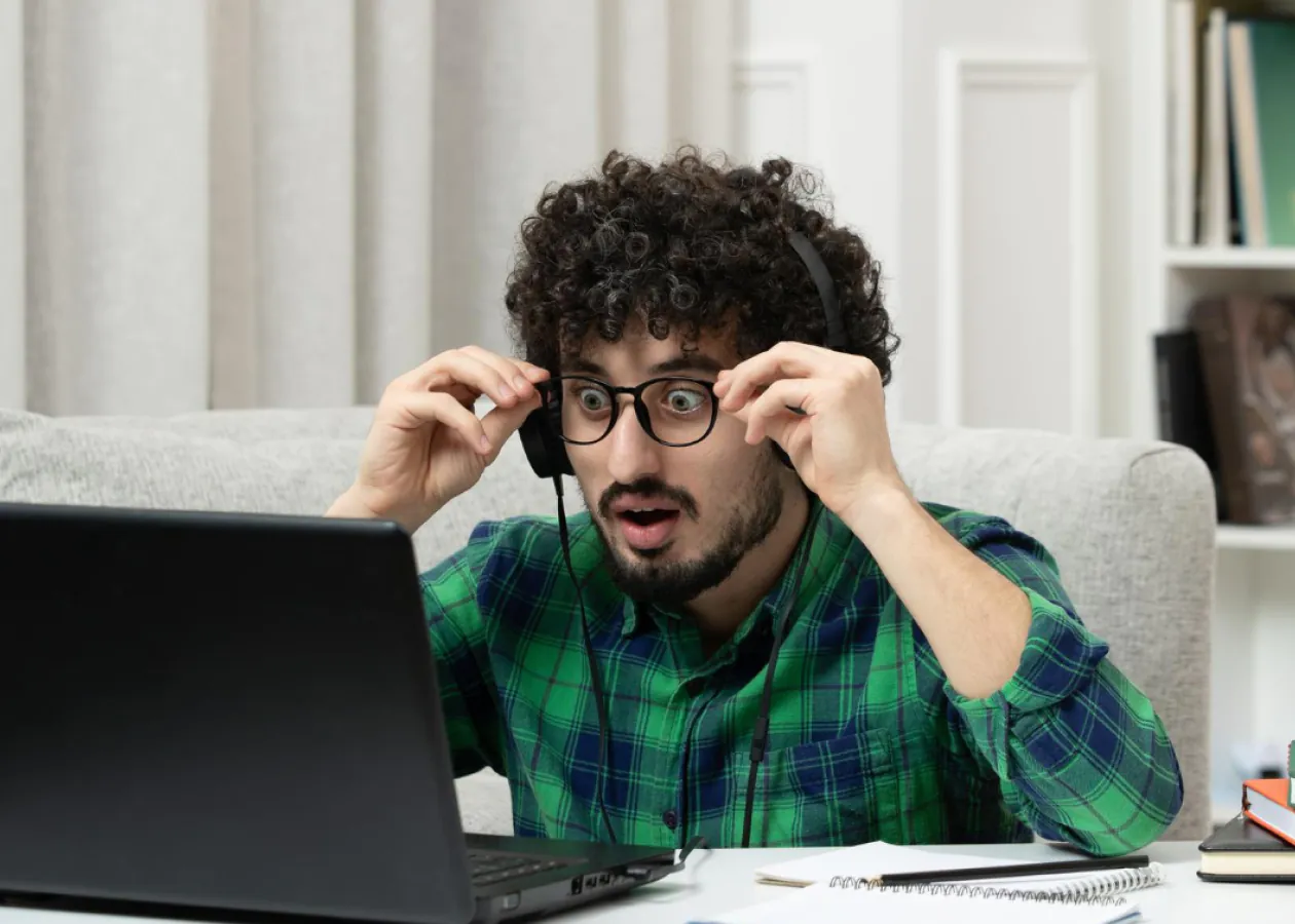 A guy wearing headphones, sitting in front of a computer, with a surprised expression on his face.